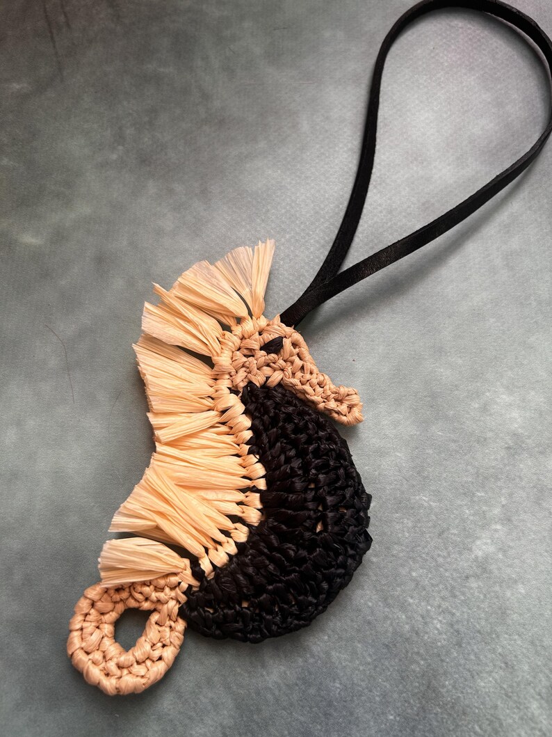May include: A seahorse-shaped ornament crafted from woven raffia in natural beige and black. The seahorse features a fringed beige dorsal fin, a solid black body, and a curled tail. A thin black cord is attached to the top for hanging purposes.