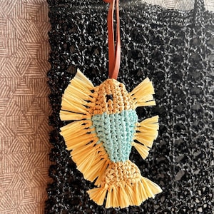 May include: A macrame fish ornament featuring a light blue body and tan raffia-like fringe fins and tail. The fish is suspended by a brown leather cord and hangs in front of a black woven tote bag with a textured, open-grid pattern design.