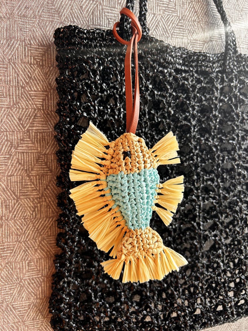 May include: A macrame fish ornament featuring a light blue body and tan raffia-like fringe fins and tail. The fish is suspended by a brown leather cord and hangs in front of a black woven tote bag with a textured, open-grid pattern design.