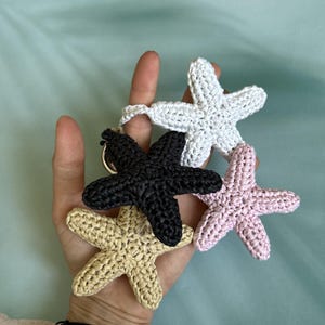 May include: A set of four crocheted starfish keychains in white, black, light pink, and beige. Each starfish features a textured, knotted yarn construction and is attached to a circular metal ring, creating a cluster of marine-themed accessories.