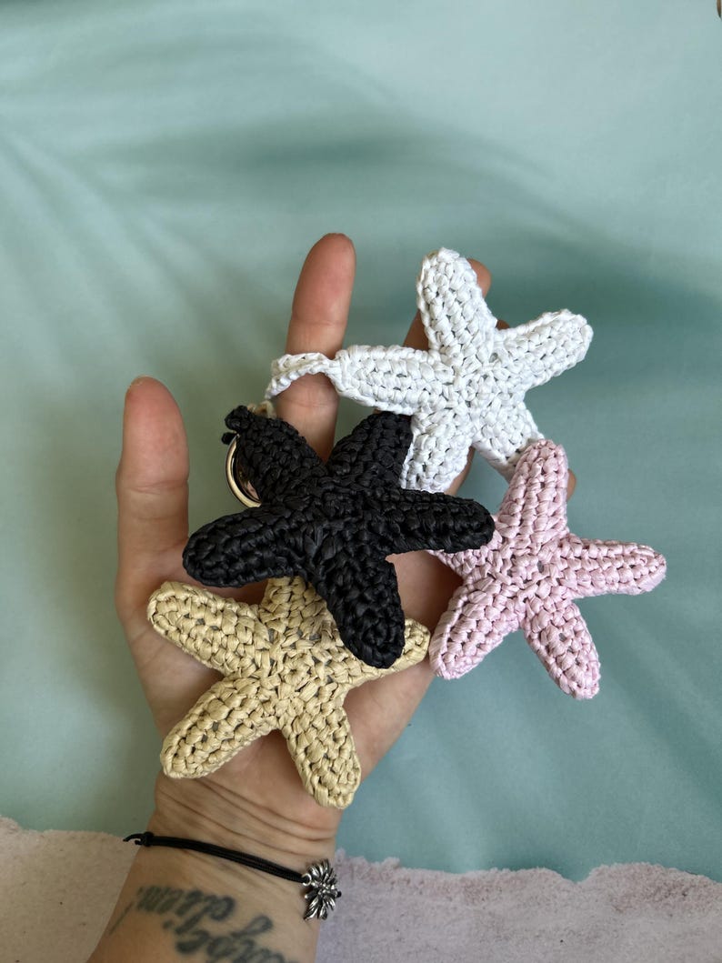 May include: A set of four crocheted starfish keychains in white, black, light pink, and beige. Each starfish features a textured, knotted yarn construction and is attached to a circular metal ring, creating a cluster of marine-themed accessories.