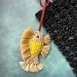 May include: A fish-shaped charm crafted from beige and yellow cord with fringed fins and tail, featuring a small black bead eye. The charm is attached to a thin, reddish-brown leather strap, which is looped through a black crocheted mesh bag.