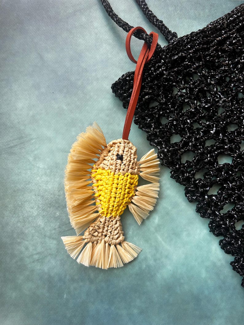 May include: A fish-shaped charm crafted from beige and yellow cord with fringed fins and tail, featuring a small black bead eye. The charm is attached to a thin, reddish-brown leather strap, which is looped through a black crocheted mesh bag.