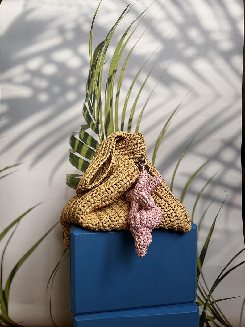 Puede incluir: Un bolso tejido de color mostaza con un charm de ganchillo rosa a juego. El bolso est&aacute; sobre un bloque azul, con hojas verdes en el fondo. El bolso tiene una parte superior doblada.