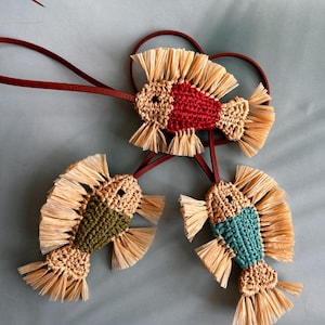 May include: Three fish-shaped ornaments crafted from woven natural-colored raffia and colored yarn in red, olive green, and teal. Each fish features fringed raffia fins and tails, a small black eye, and is attached to a thin dark brown leather cord.
