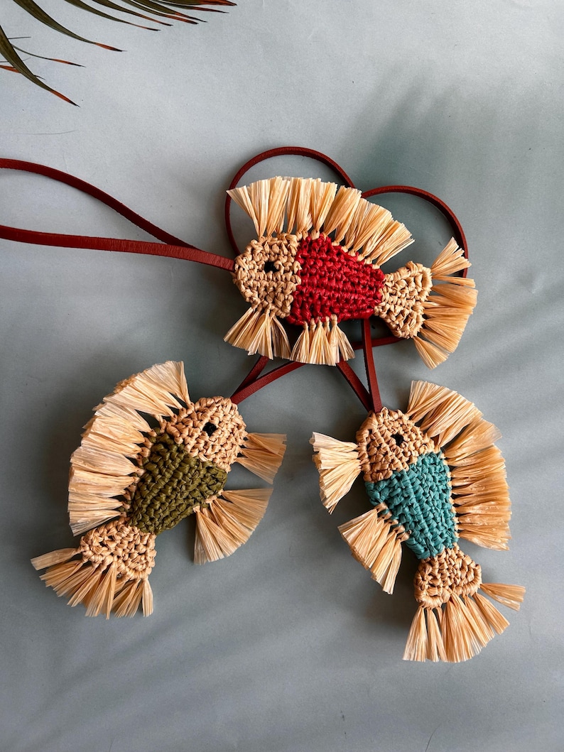 May include: Three fish-shaped ornaments crafted from woven natural-colored raffia and colored yarn in red, olive green, and teal. Each fish features fringed raffia fins and tails, a small black eye, and is attached to a thin dark brown leather cord.