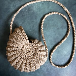 Puede incluir: Un bolso bandolera de rafia beige tejido con un diseño en espiral. El bolso tiene forma redondeada y una larga correa trenzada. El fondo es de color verde azulado apagado.