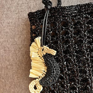 May include: A black crocheted tote bag with a textured, open-weave pattern and matching black handles. Attached to the handle is a seahorse-shaped charm crafted from black crocheted yarn and light beige raffia-like material with a black leather cord.