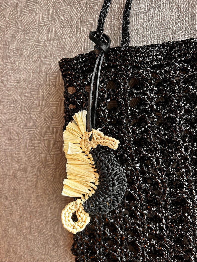 May include: A black crocheted tote bag with a textured, open-weave pattern and matching black handles. Attached to the handle is a seahorse-shaped charm crafted from black crocheted yarn and light beige raffia-like material with a black leather cord.