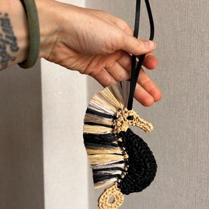 May include: A seahorse-shaped ornament crafted from woven raffia and cord. The body features a textured black crocheted section, while the dorsal fin is composed of layered, fan-like strips in alternating shades of metallic silver, black, and tan.