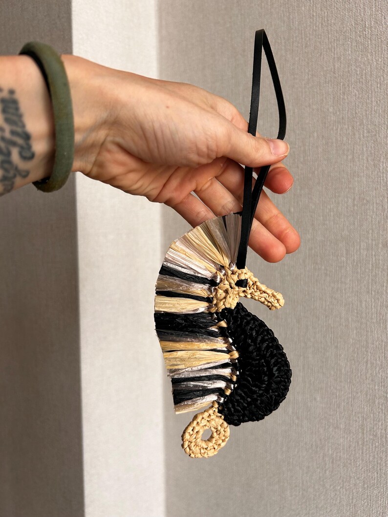 May include: A seahorse-shaped ornament crafted from woven raffia and cord. The body features a textured black crocheted section, while the dorsal fin is composed of layered, fan-like strips in alternating shades of metallic silver, black, and tan.