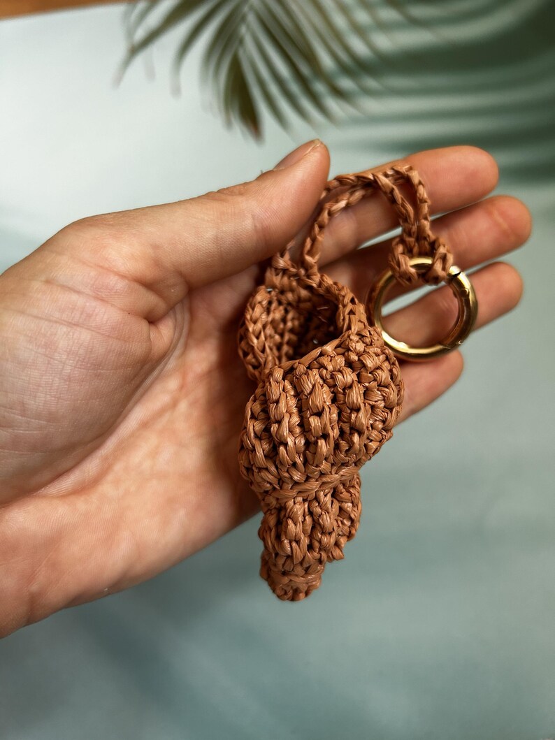 May include: A woven, seashell-shaped keychain in a warm brown hue. The keychain features a gold-colored ring for attaching keys. The seashell design is intricately woven, showcasing a textured surface. The background is a soft, muted blue.