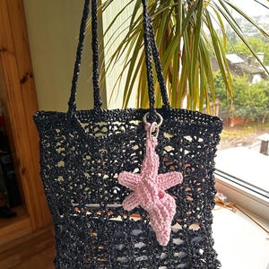 Puede incluir: Bolso tote negro de crochet con asas largas. Un charm de estrella de mar rosa de crochet cuelga de la bolsa. La bolsa tiene un dise&ntilde;o de tejido abierto, lo que permite ver el contenido en el interior. La bolsa est&aacute; sujeta por una mano.