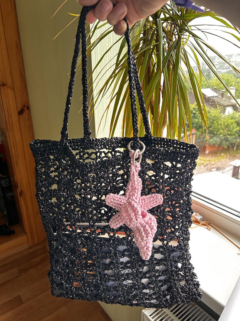 Puede incluir: Bolso tote negro de crochet con asas largas. Un charm de estrella de mar rosa de crochet cuelga de la bolsa. La bolsa tiene un dise&ntilde;o de tejido abierto, lo que permite ver el contenido en el interior. La bolsa est&aacute; sujeta por una mano.