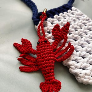 May include: A bright red crocheted lobster charm with small black bead eyes, attached by a gold-toned metal ring to a navy blue and white crocheted pouch featuring a thick navy blue strap and a textured, woven stitch pattern throughout.