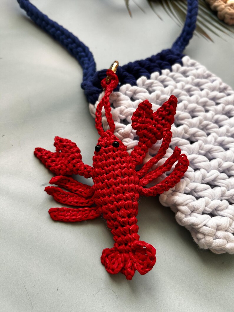 May include: A bright red crocheted lobster charm with small black bead eyes, attached by a gold-toned metal ring to a navy blue and white crocheted pouch featuring a thick navy blue strap and a textured, woven stitch pattern throughout.