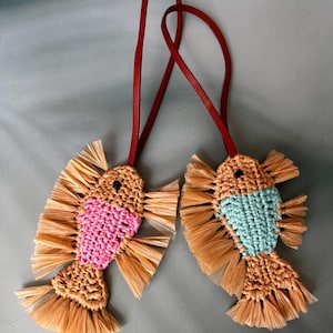 May include: Two fish-shaped ornaments crafted from tan raffia-like material, each featuring a long red leather cord for hanging. One fish has a pink woven center, while the other has a light blue woven center, both with fringed fins and tails.