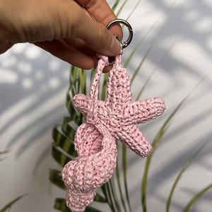 Puede incluir: Un llavero rosa tejido a ganchillo con una estrella de mar y una concha. El llavero est&aacute; unido a un anillo de metal plateado. El fondo incluye follaje verde y blanco.