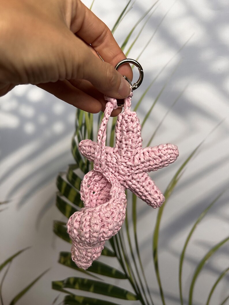 Puede incluir: Un llavero rosa tejido a ganchillo con una estrella de mar y una concha. El llavero est&aacute; unido a un anillo de metal plateado. El fondo incluye follaje verde y blanco.
