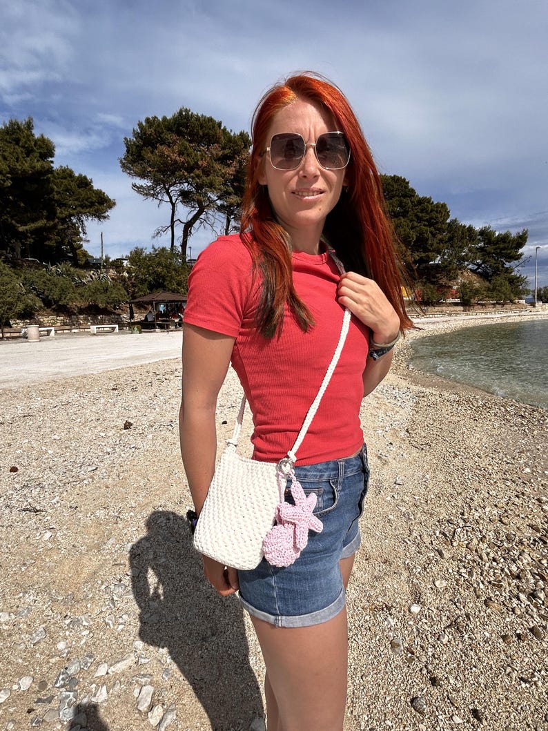 May include: A small, off-white crocheted crossbody bag featuring a textured, woven pattern and a long, thin matching strap. A light pink, crocheted starfish-shaped charm is attached to the silver-toned metal ring on the side of the bag.