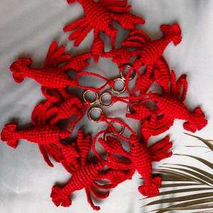 May include: A collection of six bright red crocheted lobster keychains arranged in a circular pattern. Each lobster features a textured yarn body, small black bead eyes, and is attached to a circular gold-toned metal keyring by a short cord.