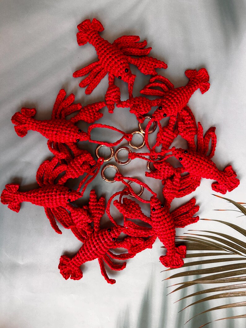 May include: A collection of six bright red crocheted lobster keychains arranged in a circular pattern. Each lobster features a textured yarn body, small black bead eyes, and is attached to a circular gold-toned metal keyring by a short cord.