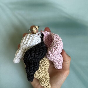 Puede incluir: Llaveros artesanales con forma de concha marina en blanco, negro, beige y rosa. Cada llavero est&aacute; hecho de material tejido y se sujeta a un llavero plateado. Los llaveros se sostienen en una mano sobre un fondo azul claro. Ideal para regalos.