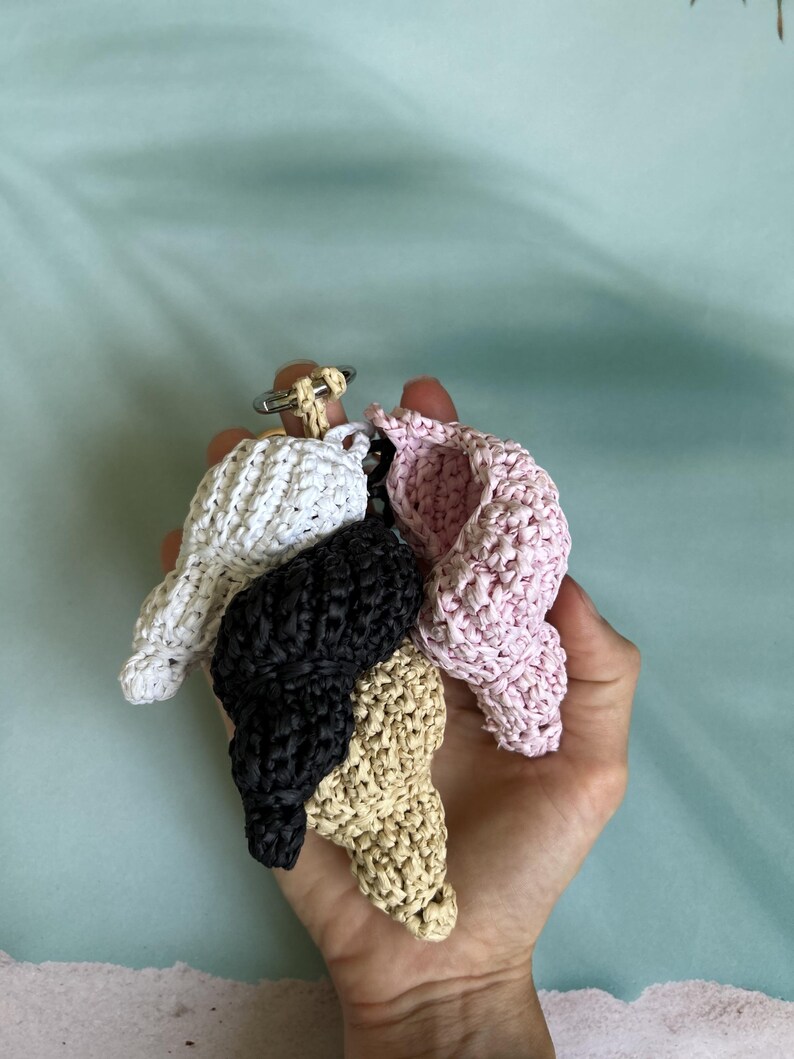 Puede incluir: Llaveros artesanales con forma de concha marina en blanco, negro, beige y rosa. Cada llavero est&aacute; hecho de material tejido y se sujeta a un llavero plateado. Los llaveros se sostienen en una mano sobre un fondo azul claro. Ideal para regalos.
