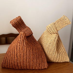 Könnte beinhalten: Zwei gewebte Strohhandtaschen. Eine ist warmbraun, die andere hellbeige. Beide haben einen geknoteten Griff und eine dreieckige Form. Die Taschen stehen auf einer Holzoberfläche.