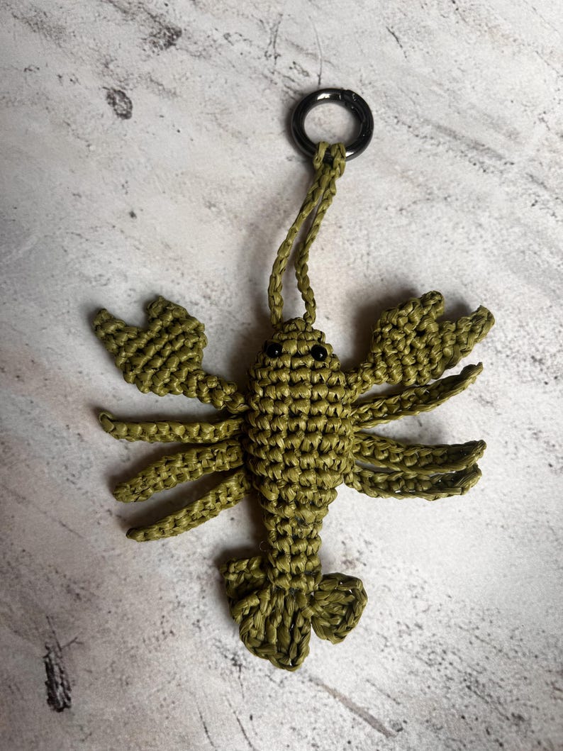 May include: An olive green crocheted lobster keychain featuring a textured body, two large front claws, multiple legs, and a fan-shaped tail. The lobster has two small black bead eyes and is attached to a circular silver-toned metal ring.