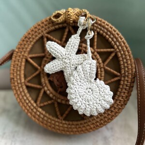 May include: A round, woven brown rattan bag featuring a star-shaped and a spiral shell-shaped white crochet charm attached to the top. The bag has a brown leather strap and a central star pattern woven into the front of the circular body.