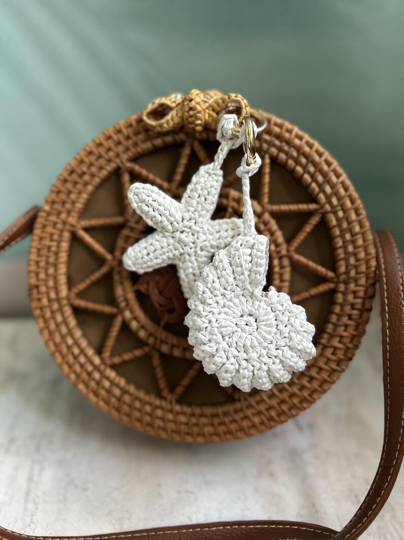 May include: A round, woven brown rattan bag featuring a star-shaped and a spiral shell-shaped white crochet charm attached to the top. The bag has a brown leather strap and a central star pattern woven into the front of the circular body.