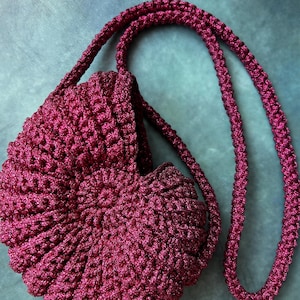 Pode incluir: Uma bolsa de crochê redonda em tom magenta profundo com um padrão texturizado em espiral que lembra uma concha. A bolsa inclui uma alça de ombro longa e correspondente em crochê com um ponto denso e consistente em toda a peça.