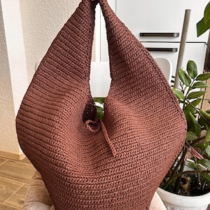 May include: A dark brown, solid-colored hobo bag crafted with a dense, textured crochet stitch. The bag features a wide, integrated shoulder strap that forms a triangular silhouette, with a small drawstring closure detail at the center opening.