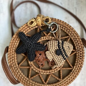 May include: A round woven rattan crossbody bag with a brown leather strap. The front features a crocheted black starfish charm and a beige crocheted fish charm with black accents attached to a silver metal ring at the top of the bag.