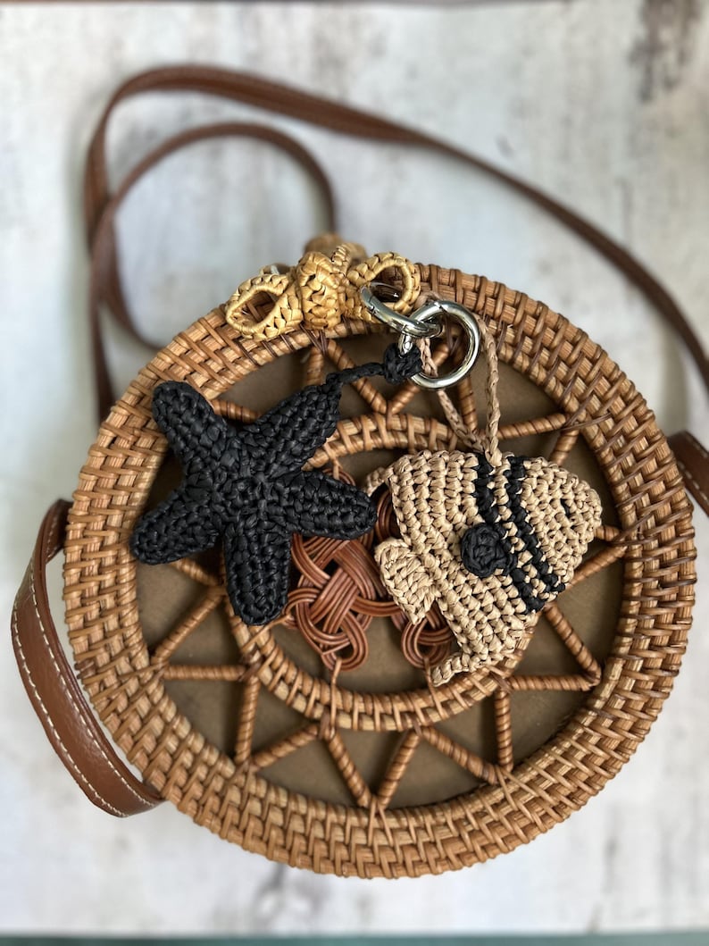 May include: A round woven rattan crossbody bag with a brown leather strap. The front features a crocheted black starfish charm and a beige crocheted fish charm with black accents attached to a silver metal ring at the top of the bag.