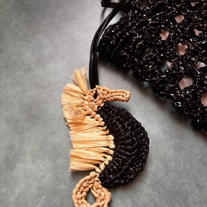 May include: A seahorse-shaped bag charm featuring a black crocheted body and a light beige raffia-like fringe along the back. The seahorse has a coiled tail and a snout made of light beige yarn, attached to a black strap near a black crocheted bag.