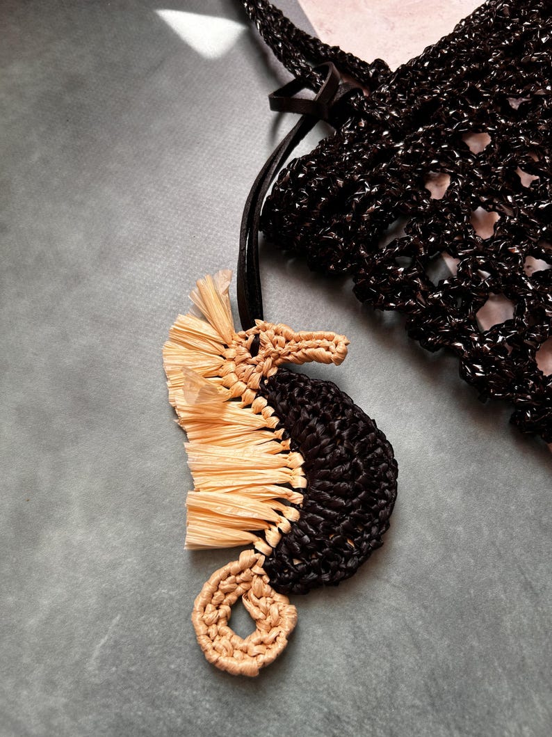 May include: A seahorse-shaped bag charm featuring a black crocheted body and a light beige raffia-like fringe along the back. The seahorse has a coiled tail and a snout made of light beige yarn, attached to a black strap near a black crocheted bag.