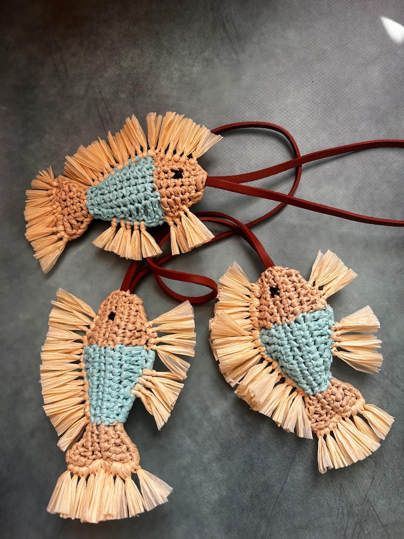 May include: Three fish-shaped ornaments crafted from light blue and beige crocheted yarn, featuring fringed beige raffia fins and tails. Each fish is attached to a thin, dark brown leather cord for hanging, with a single black bead for the eye.