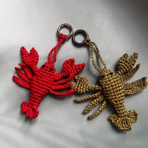 May include: Two handmade crochet lobster keychains, one in bright red and one in olive green. Each lobster features small black bead eyes, detailed pincers, and multiple legs, attached to a circular metal keyring for easy attachment to bags or keys.