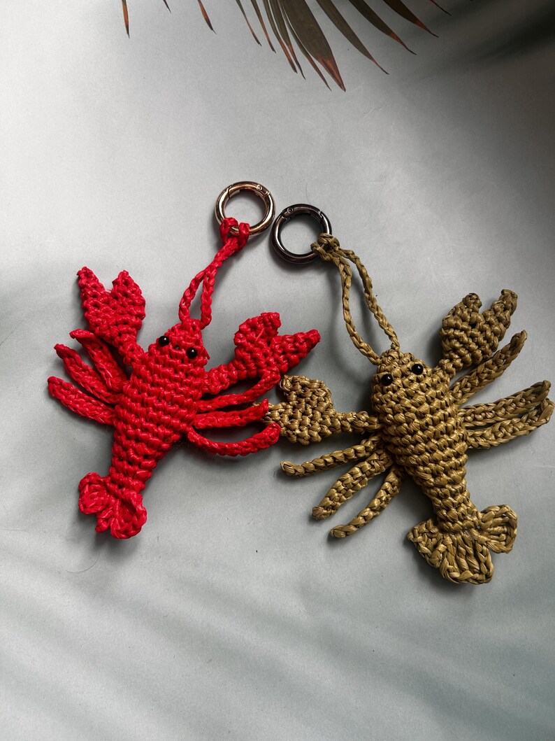 May include: Two handmade crochet lobster keychains, one in bright red and one in olive green. Each lobster features small black bead eyes, detailed pincers, and multiple legs, attached to a circular metal keyring for easy attachment to bags or keys.