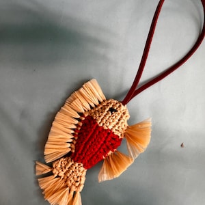 May include: A fish-shaped ornament crafted from woven beige and red cord, featuring fringed beige fins and a tail. A dark red leather loop is attached to the top for hanging, providing a contrast to the textured, multi-colored fiber body.