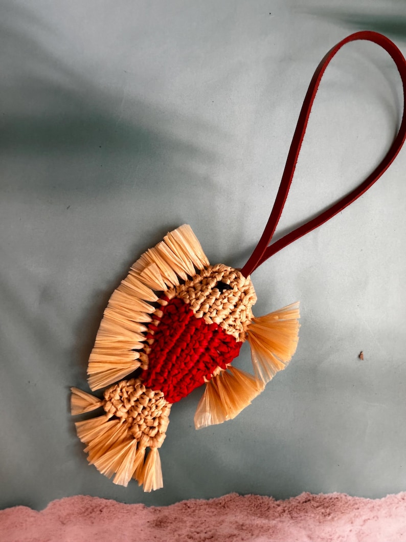 May include: A fish-shaped ornament crafted from woven beige and red cord, featuring fringed beige fins and a tail. A dark red leather loop is attached to the top for hanging, providing a contrast to the textured, multi-colored fiber body.