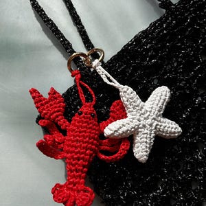 May include: A black crocheted bag featuring two attached charms: a bright red crocheted lobster and a white crocheted starfish. The charms are connected to the bag by a gold-toned metal ring, and the bag has a black crocheted strap.