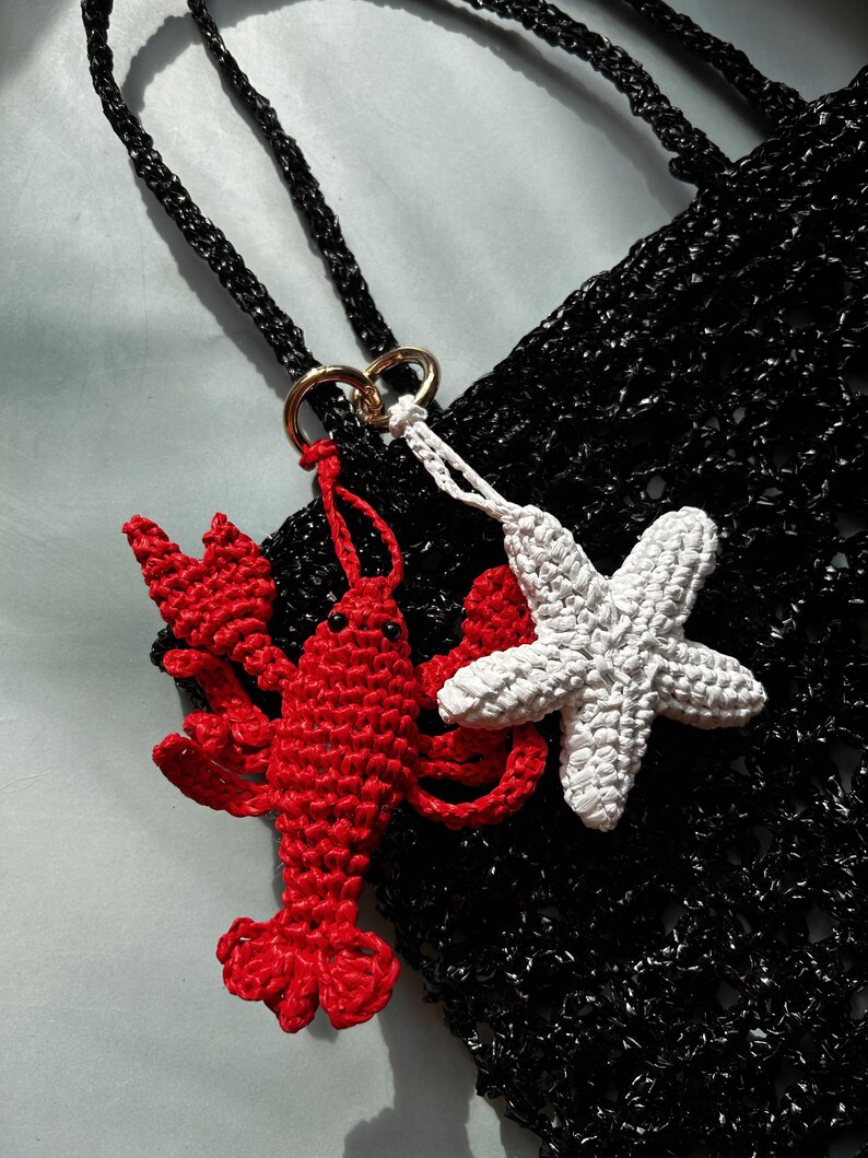 May include: A black crocheted bag featuring two attached charms: a bright red crocheted lobster and a white crocheted starfish. The charms are connected to the bag by a gold-toned metal ring, and the bag has a black crocheted strap.