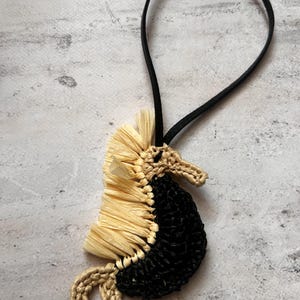 May include: A seahorse-shaped ornament crafted from black and beige cord, featuring a textured beige raffia-like mane along the back and a coiled tail. A thin black elastic loop is attached to the top for hanging this decorative piece.