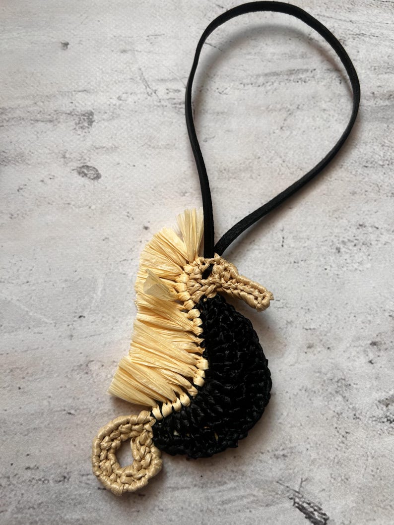 May include: A seahorse-shaped ornament crafted from black and beige cord, featuring a textured beige raffia-like mane along the back and a coiled tail. A thin black elastic loop is attached to the top for hanging this decorative piece.