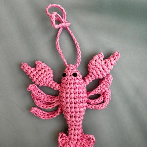 May include: A pink crocheted lobster ornament featuring a textured, knotted yarn construction, two black bead eyes, multiple articulated legs, large front claws, and a looped string at the top for hanging, crafted in a vibrant solid pink color.