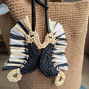 May include: A beige crocheted tote bag featuring a hanging charm composed of two seahorse-shaped figures. The seahorses are crafted from black yarn and light-colored raffia-like fringe, suspended by a black leather cord attached to the bag handle.