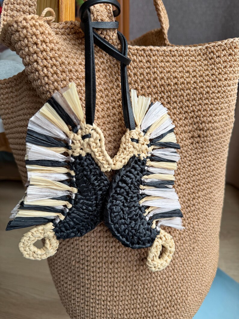 May include: A beige crocheted tote bag featuring a hanging charm composed of two seahorse-shaped figures. The seahorses are crafted from black yarn and light-colored raffia-like fringe, suspended by a black leather cord attached to the bag handle.